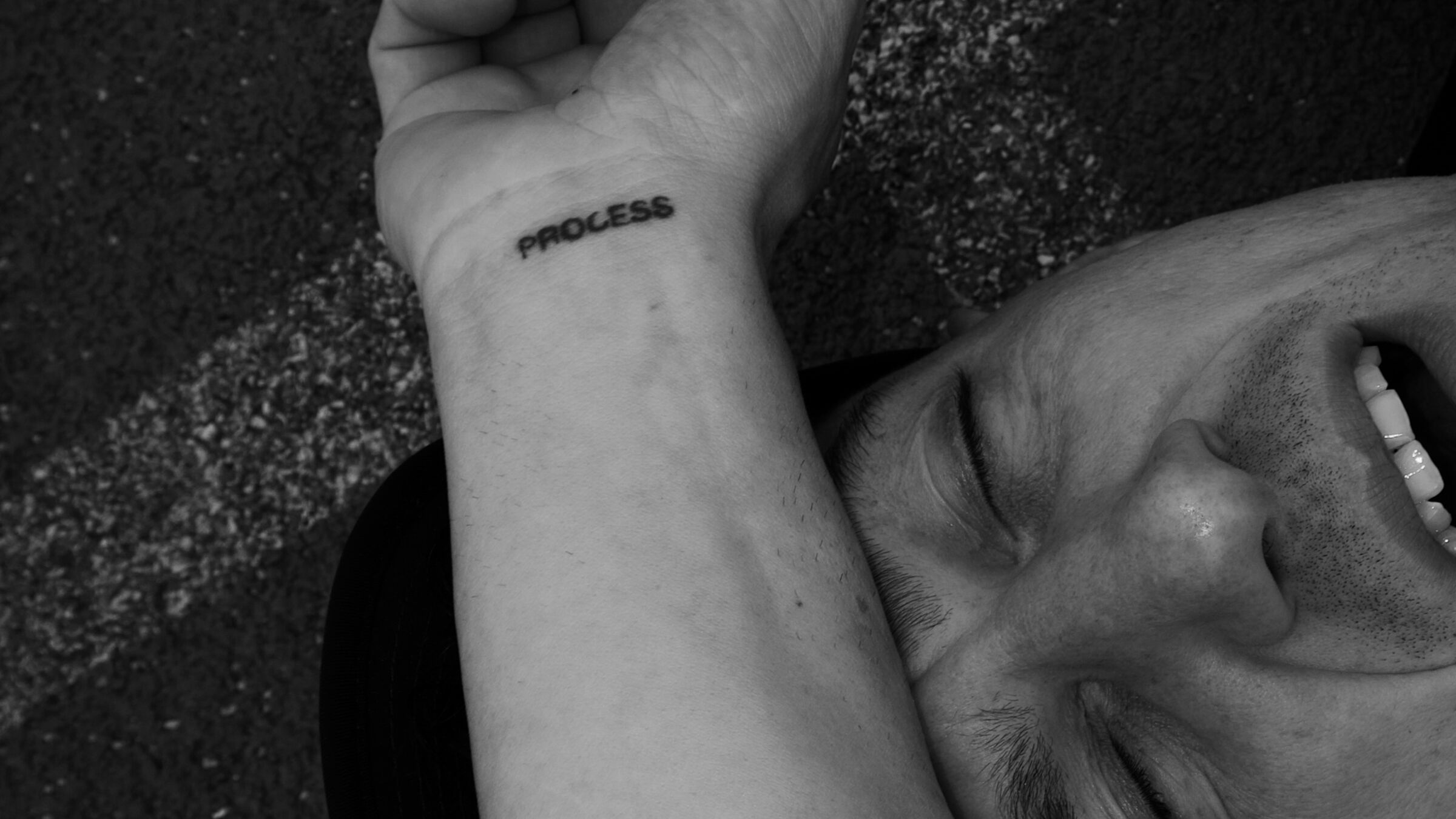 An athlete lying on asphalt, laughing, with a PROCESS tattoo on the wrist