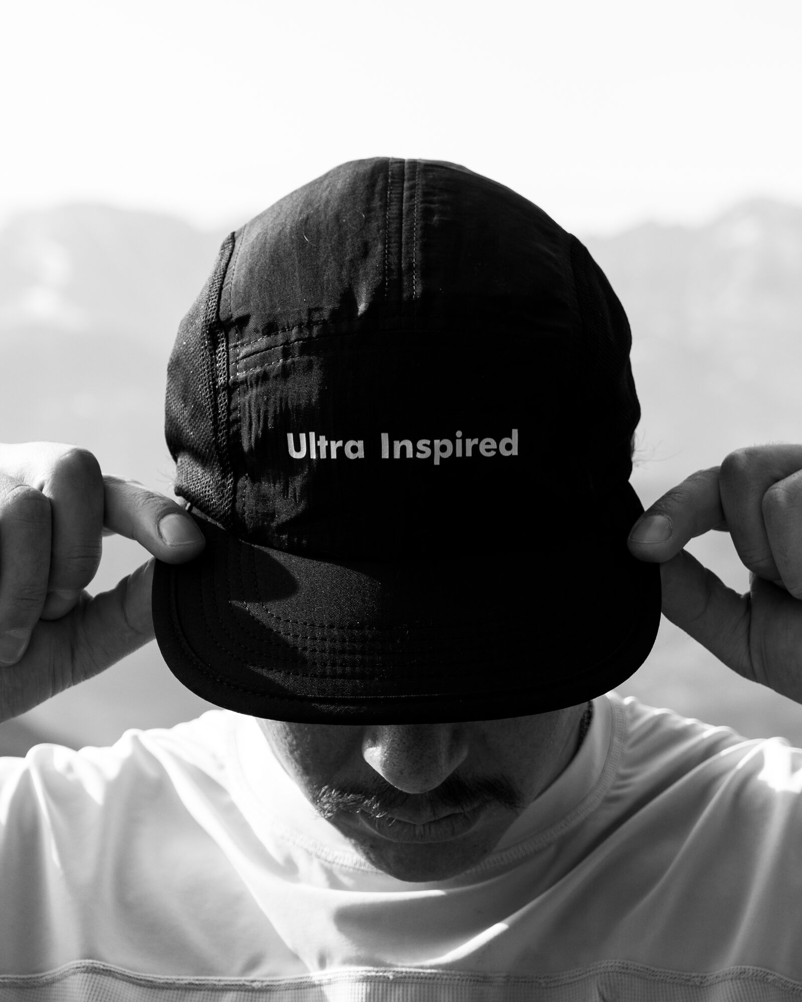 An athlete pulling down a black Ultra Inspired cap, mountains behind
